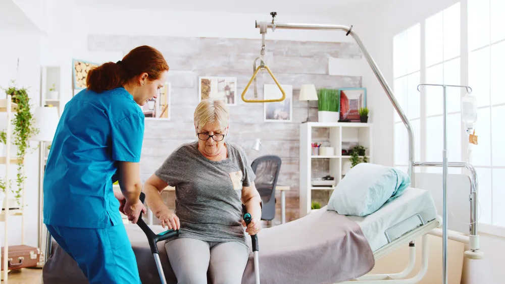 Zorgmedewerker in blauw uniform helpt een oudere vrouw met krukken overeind in een woonkamer